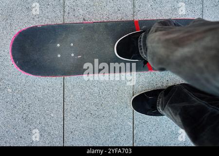 Skateboarder in sella allo skateboard. Vista dall'alto dei piedi adolescenti che indossano jeans e sneakers nere da skateboard sulla strada. Attività ricreative ed extra Foto Stock