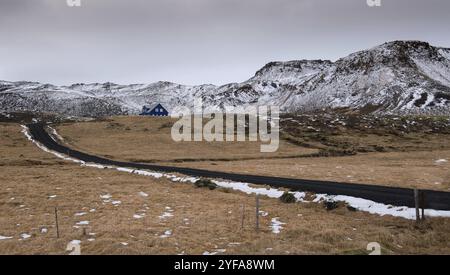 Tipico paesaggio islandese nella penisola di Reykjanes con montagne coperte di neve e strade asfaltate vuote che conducono ad una casa di villeggiatura blu Foto Stock