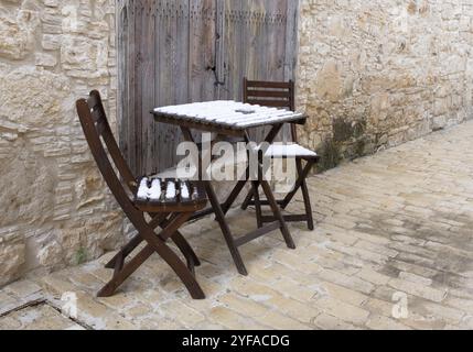 Sedie di legno coperte di neve vuote e tavolino da caffè su un sentiero in pietra. Vecchio villaggio lofou quartiere cipro Foto Stock