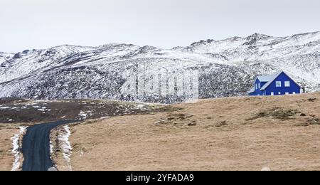 Tipico paesaggio islandese nella penisola di Reykjanes con montagne coperte di neve e strade asfaltate vuote che conducono ad una casa di villeggiatura blu Foto Stock