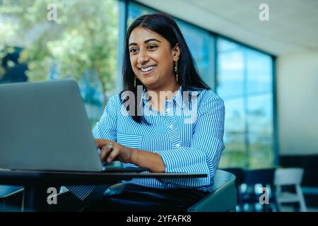 Una donna d'affari indiana è impegnata in una riunione virtuale, sorridendo mentre lavora sul suo portatile. L'ambiente luminoso e il suo comportamento positivo suggeriscono Foto Stock