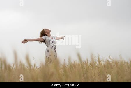 Donna felice con un abito floreale si erge in mezzo al grano dorato, godendosi il paesaggio sereno Foto Stock