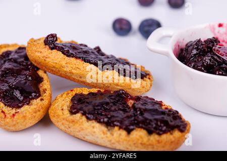 Una fetta di pane tostato con marmellata di mirtilli si trova accanto a un vaso di marmellata su sfondo bianco. Fresca e vivace, questa colazione è ricca di sapori Foto Stock