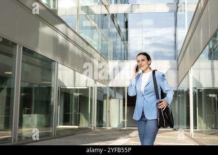 Una donna dirigente sta camminando fuori di fronte al suo moderno edificio per uffici mentre parla al suo cellulare Foto Stock