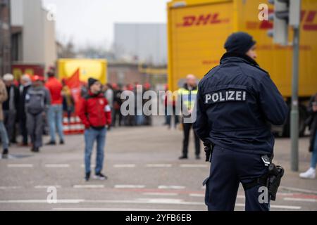 Augusta, Baviera, Germania - 4 novembre 2024: Agente di polizia della polizia bavarese in Svevia di Augusta, blocca la strada per lo sciopero, dietro il camion DHL o il corriere DHL, durante lo sciopero di avvertimento di MAN IG Metall. *** Polizist bzw. Polizeibeamter der bayrischen Polizei ad Augusta Schwaben, sperrt Straße für Streik, dahinter DHL LKW bzw. DHL Paketfahrzeug, beim IG Metall Warnstreik der MAN. Foto Stock