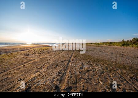 Un'ampia spiaggia di sabbia che si estende in lontananza con il sole che tramonta sul mare, lasciando toni caldi sul terreno e un vasto cielo aperto sopra. Foto Stock