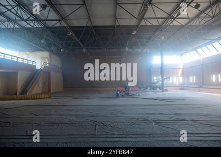 Palestra in fase di ristrutturazione con attrezzature da costruzione, pavimenti coperti e travi visibili, luce del sole che splende attraverso le alte finestre. Foto Stock