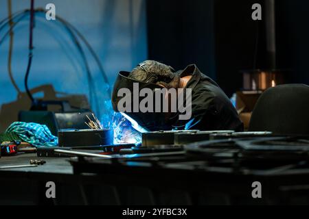 Saldatrice che lavora intensamente su un progetto metallico, scintille che volano dalla torcia di saldatura illuminando l'officina poco illuminata. Foto Stock