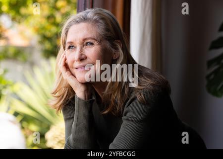 Donna anziana sorridente che guarda fuori dalla finestra, godendo momenti tranquilli a casa Foto Stock