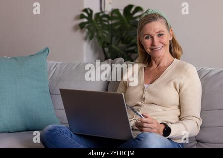 donna anziana che usa il laptop sul divano a casa, sorride e tiene gli occhiali Foto Stock