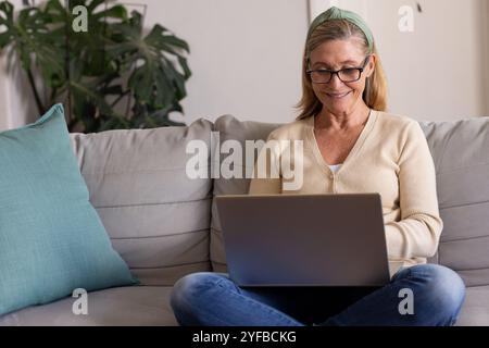 Donna anziana sorridente che utilizza il laptop su un comodo divano a casa, si sente rilassata Foto Stock