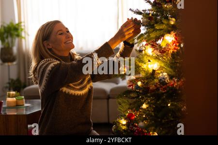 Donna anziana sorridente che decora l'albero di Natale con ornamenti a casa Foto Stock