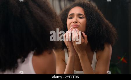 Preoccupata giovane bella donna afro-americana triste ragazza etnica che guarda in specchio stressato per le rughe facciali problema acne frustrato turbato toccando Foto Stock