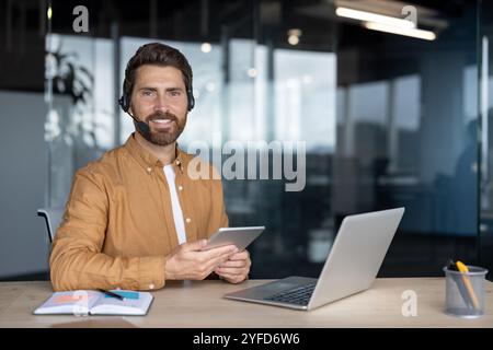 Uomo d'affari maturo e sicuro che utilizza tablet e notebook in un ufficio moderno. Auricolare per comunicazioni professionali. Sorridente, coinvolgente, che riflette l'ambiente di lavoro produttivo e l'uso della tecnologia. Foto Stock