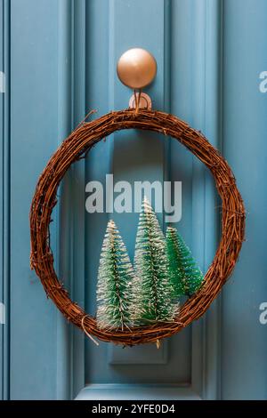 Una corona di Natale decorativa con piccoli alberi smerigliati è appesa su una porta blu, evocando un'atmosfera accogliente e festosa Foto Stock