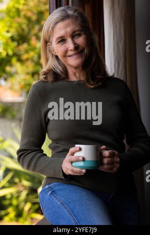 Una donna anziana sorridente che si gusta un caffè vicino alla finestra a casa, si sente rilassata Foto Stock