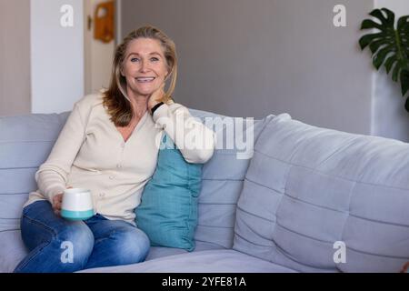 Donna anziana sorridente che si rilassa sul divano a casa, tenendo una tazza di caffè Foto Stock