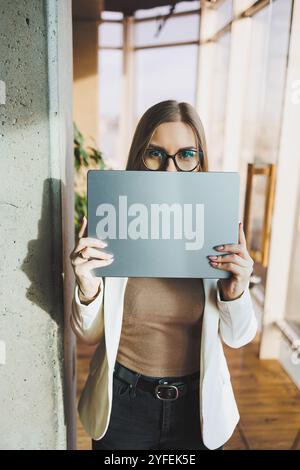 Una giovane donna d'affari con lunghi capelli biondi in una giacca bianca e occhiali si regge con un laptop in mano in un ufficio. Lavora in un ambiente moderno Foto Stock