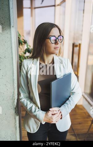 Una giovane donna d'affari con lunghi capelli biondi in una giacca bianca e occhiali si regge con un laptop in mano in un ufficio. Lavora in un ambiente moderno Foto Stock