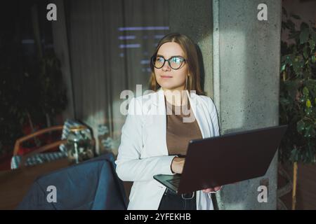 Una giovane donna d'affari con lunghi capelli biondi in una giacca bianca e occhiali si regge con un laptop in mano in un ufficio. Lavora in un ambiente moderno Foto Stock