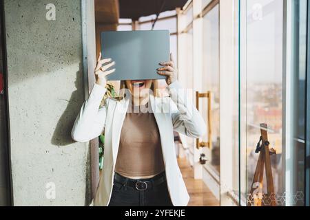 Una giovane donna d'affari con lunghi capelli biondi in una giacca bianca e occhiali si regge con un laptop in mano in un ufficio. Lavora in un ambiente moderno Foto Stock