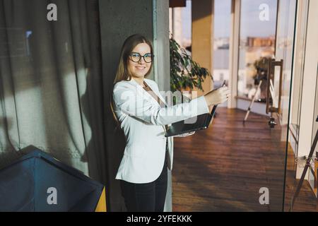 Una giovane donna d'affari con lunghi capelli biondi in una giacca bianca e occhiali si regge con un laptop in mano in un ufficio. Lavora in un ambiente moderno Foto Stock