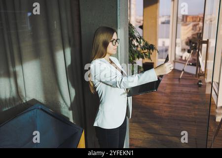 Una giovane donna d'affari con lunghi capelli biondi in una giacca bianca e occhiali si regge con un laptop in mano in un ufficio. Lavora in un ambiente moderno Foto Stock