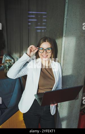 Una giovane donna d'affari con lunghi capelli biondi in una giacca bianca e occhiali si regge con un laptop in mano in un ufficio. Lavora in un ambiente moderno Foto Stock