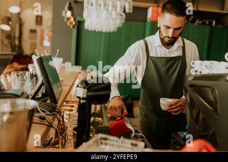 In un caffè caldo e invitante, un barista dedicato versa sapientemente il latte in una tazza da caffè, creando un'esperienza deliziosa per i clienti. Foto Stock