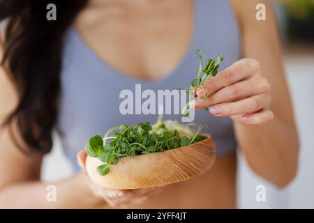 La giovane donna tiene in mano una ciotola di legno di microgreen freschi, aggiungendo un tocco salutare alla sua dieta Foto Stock