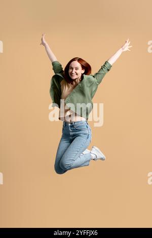 Una giovane donna allegra salta con gioia nell'aria, esprimendo pura felicità ed emozione in un solido sfondo beige Foto Stock