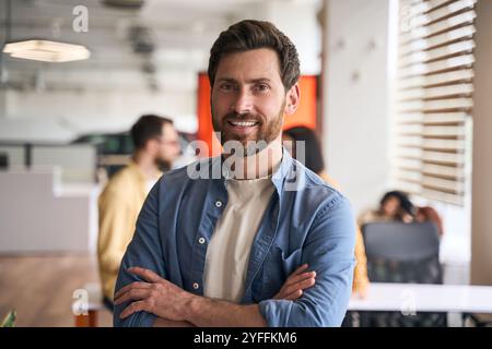 Il giovane uomo d'affari sorride con le braccia incrociate in un ufficio affollato con il suo team che lavora sullo sfondo Foto Stock