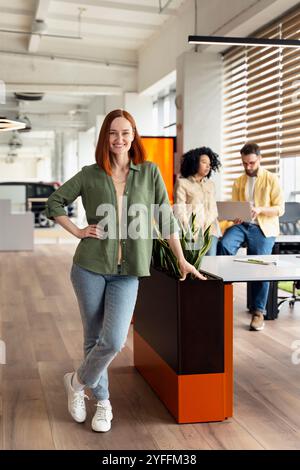Giovane donna d'affari con i capelli rossi sorridente mentre posa in un ufficio moderno con colleghi che lavorano sullo sfondo Foto Stock