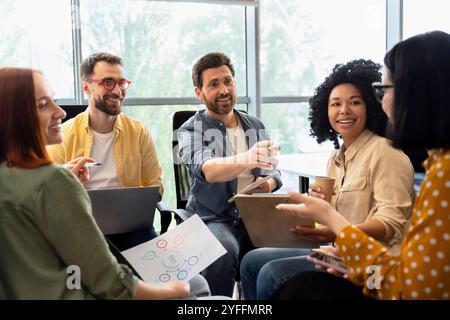 Un team di professionisti è seduto in cerchio in un ufficio moderno, discutendo un progetto e sorridendo Foto Stock