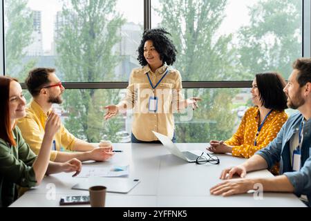 La giovane manager è in piedi e conduce una riunione di team creativi in un moderno ufficio open space, discutendo nuove idee e strategie Foto Stock