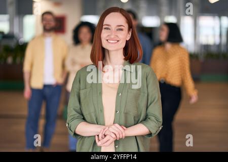 La donna d'affari sorride con le mani piegate nel corridoio di un ufficio moderno con i colleghi che camminano sullo sfondo Foto Stock