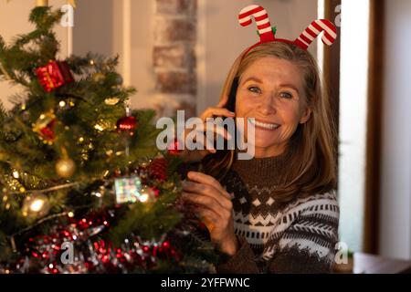 Donna anziana sorridente che decora l'albero di Natale mentre parla sullo smartphone a casa Foto Stock