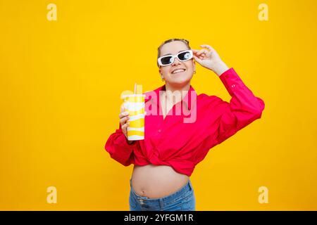 Allegra donna incinta in camicia rosa che regala una bevanda gustosa su sfondo giallo isolato. Una bevanda rinfrescante durante la gravidanza. Una donna incinta beve Foto Stock