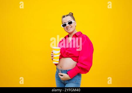 Allegra donna incinta in camicia rosa che regala una bevanda gustosa su sfondo giallo isolato. Una bevanda rinfrescante durante la gravidanza. Una donna incinta beve Foto Stock