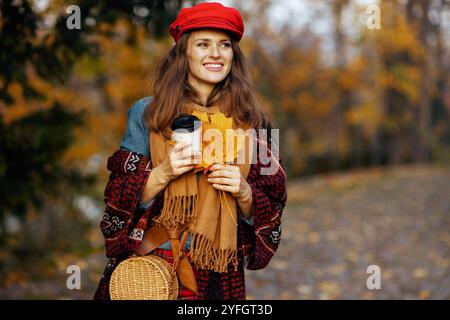 Ciao autunno. felice ed elegante donna di 40 anni in cappello rosso con foglie autunnali, sciarpa, borsa e caffè passeggiando nel parco cittadino. Foto Stock