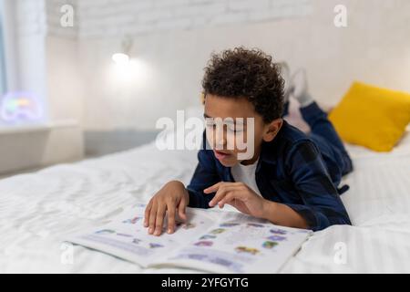 Ragazzo afroamericano dai capelli ricci sdraiato sul letto con un libro e sembra eccitato Foto Stock