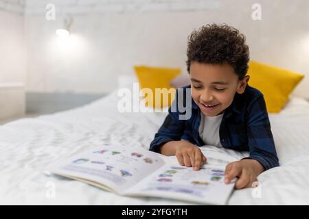 Ragazzo afroamericano dai capelli ricci sdraiato sul letto con un libro e sembra eccitato Foto Stock