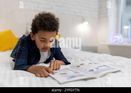 Ragazzo afroamericano dai capelli ricci sdraiato sul letto con un libro e sembra eccitato Foto Stock