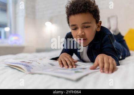 Ragazzo afroamericano dai capelli ricci sdraiato sul letto con un libro e sembra eccitato Foto Stock