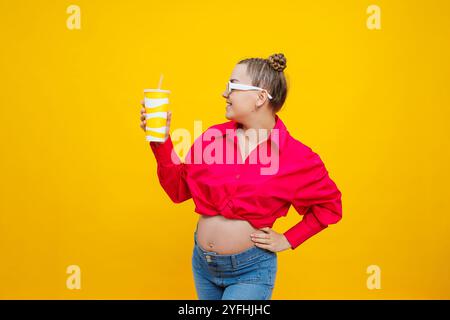 Una donna incinta in una camicia rosa beve l'acqua da una tazza di plastica. Una donna incinta su sfondo giallo con un bicchiere in mano. Wate dannoso Foto Stock