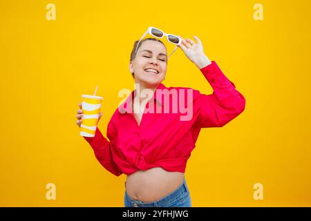 Una donna incinta in una camicia rosa beve l'acqua da una tazza di plastica. Una donna incinta su sfondo giallo con un bicchiere in mano. Wate dannoso Foto Stock