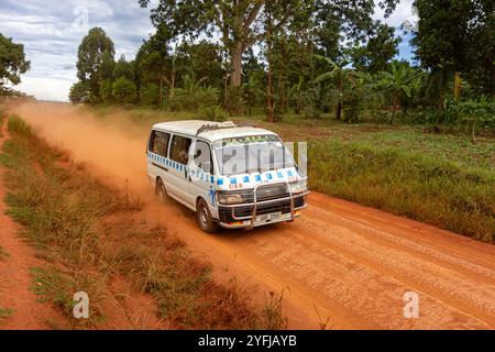 Un taxi van, noto anche come "matatu", viaggia su una strada sterrata rossastra in un'area rurale ugandese, circondata da vegetazione lussureggiante. Foto Stock