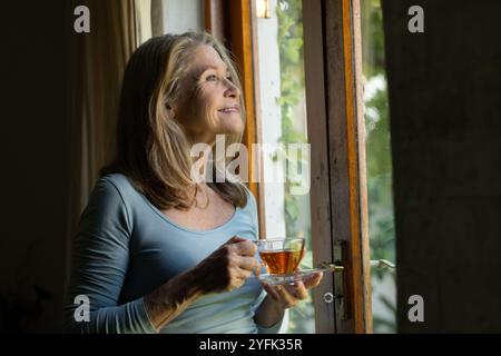 donna anziana che si gode il tè alla finestra, sorride e guarda all'esterno pacificamente, a casa Foto Stock