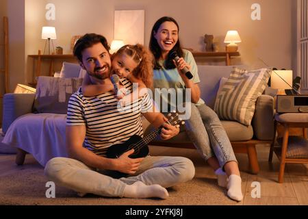 Famiglia felice con microfoni e ukulele che cantano karaoke sul divano a casa la sera Foto Stock