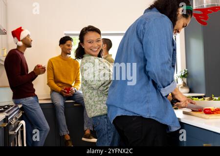 Amici multirazziali che celebrano il Natale a casa, preparano cibo e si godono momenti festivi Foto Stock